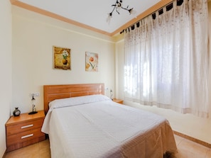 Ein Schlafzimmer mit einem hölzernen Kopfteil, einem Bett mit weißer Decke, einem Nachttisch mit einer Lampe und einem Fenster mit durchsichtigen Vorhängen.