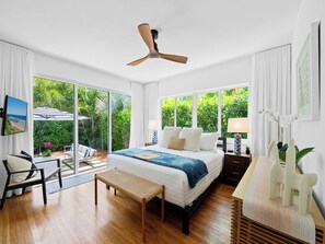 A bedroom with a bed, a ceiling fan, a TV, a bench, and a view of greenery outside. - New, Mid-Century Modern Heated Private Pool Retreat Near Beaches & Dining! (Wilton Manors)