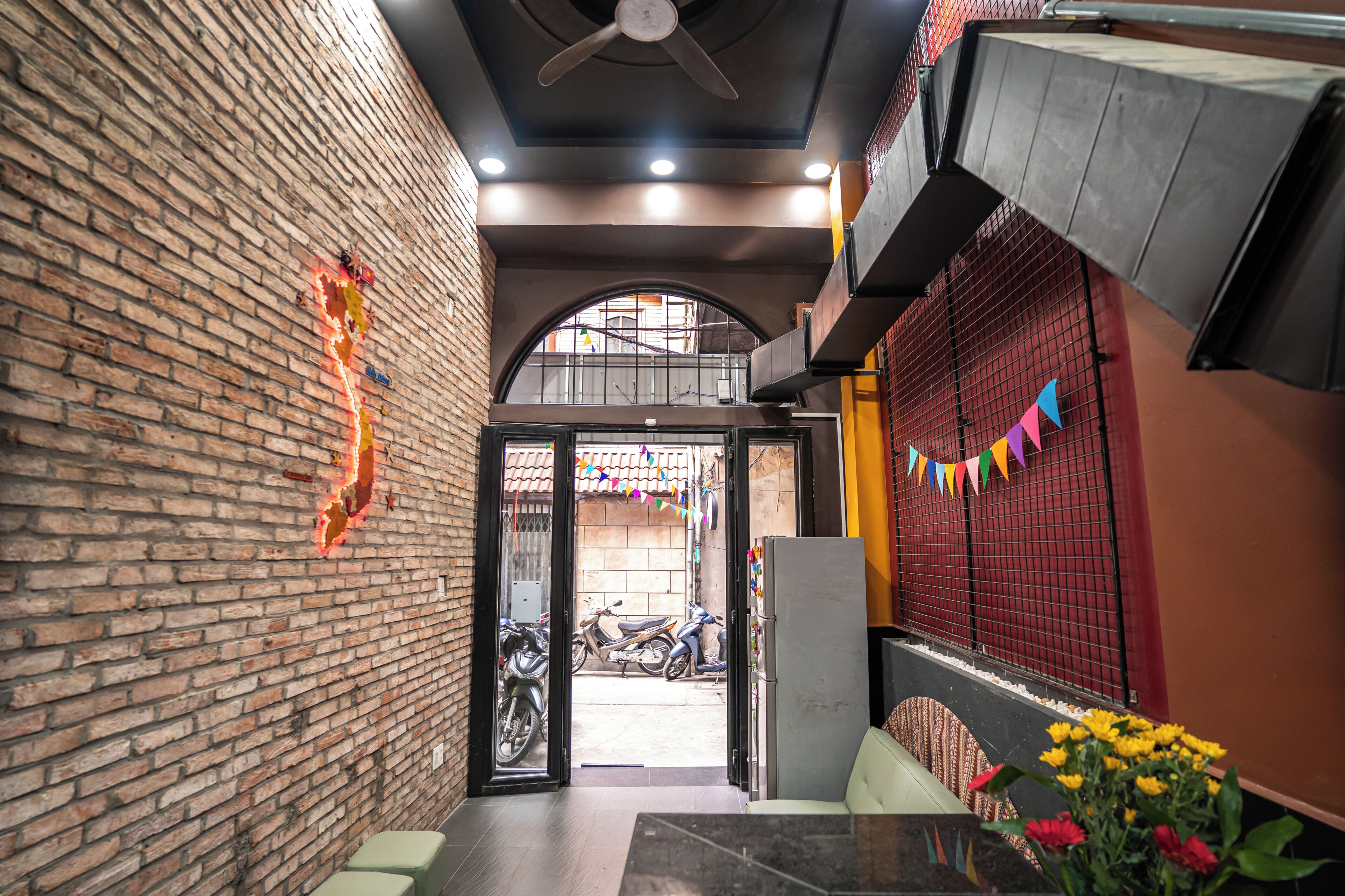 A brick-walled corridor with a ceiling fan, a table with a vase of flowers, and a view of parked scooters outside.