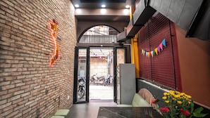 Corredor de paredes de tijolo com ventilador de teto, uma mesa com um vaso de flores e vista para scooters estacionadas lá fora.