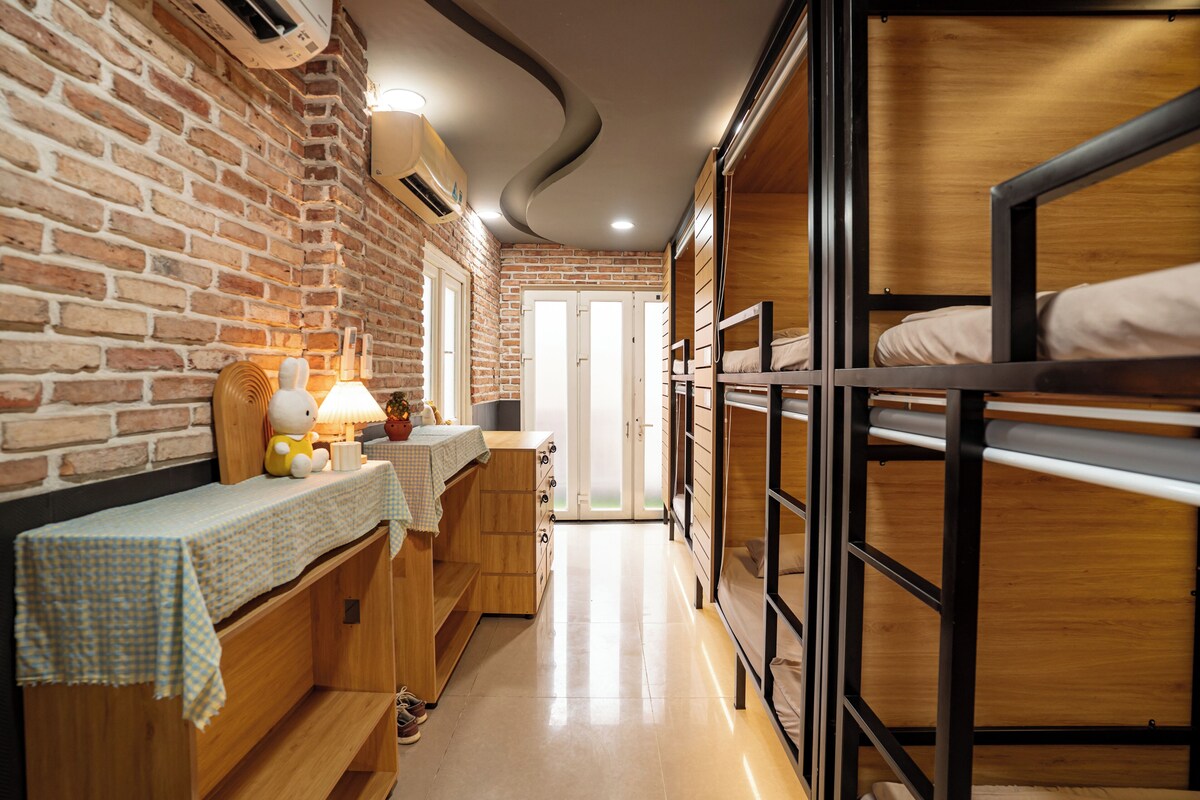 Habitación con literas, pared de ladrillo, escritorio con lámpara y ventana.