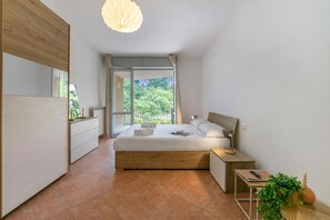A modern bedroom with a large bed, a bedside table, a nightstand, a window with a view of greenery, and a ceiling light. - Residenza il Mulino: Lake Maggiore Ispra Apt With Terrace (Ispra)