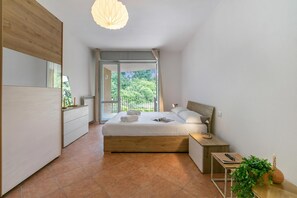 Ein modernes Schlafzimmer mit einem großen Bett, einem Nachttisch, einer Fensterbank und einer Deckenleuchte, mit Blick auf Grünflächen.