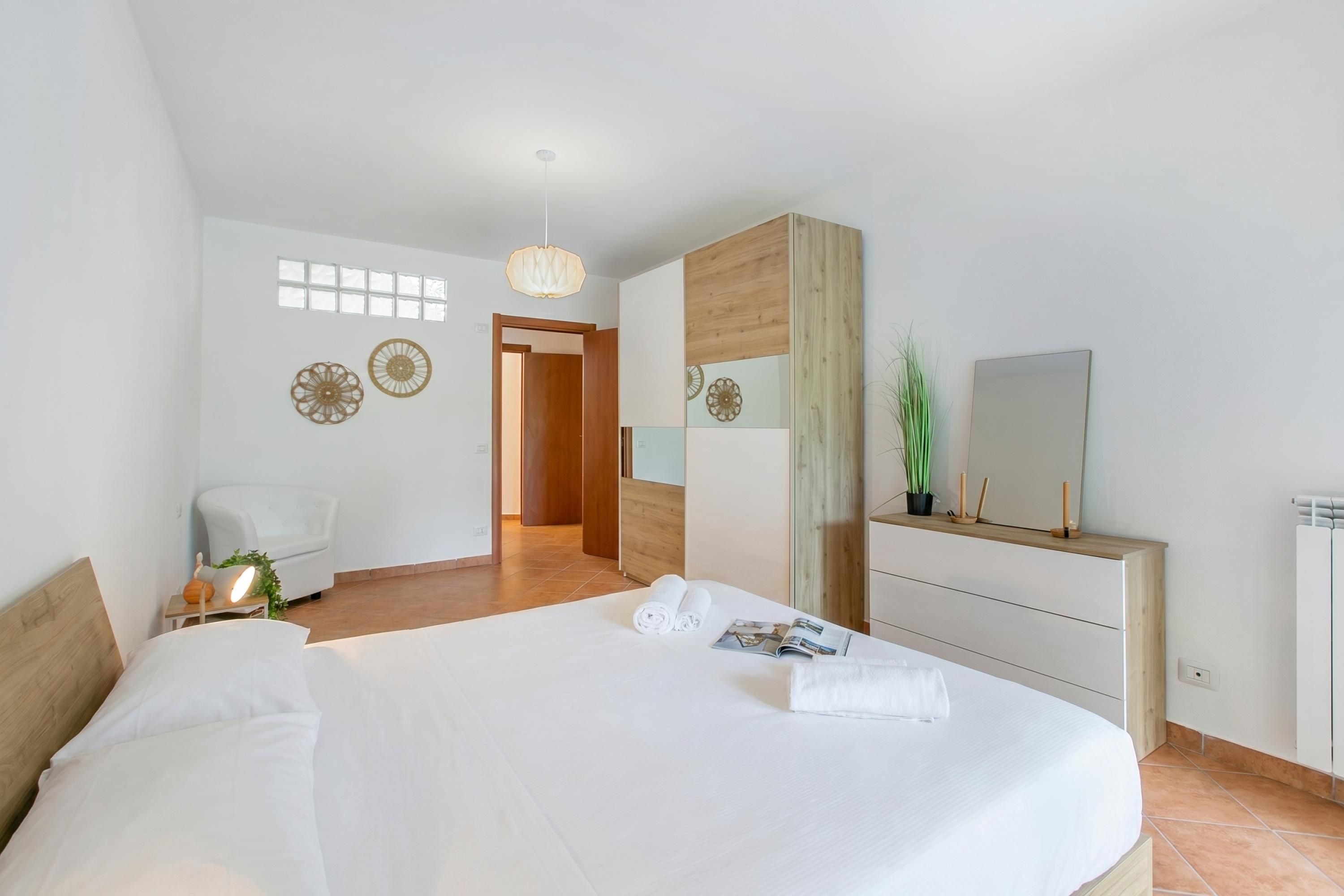 A modern hotel room with a large bed, a wooden headboard, a bedside table with a lamp, a mirror, and a dresser.