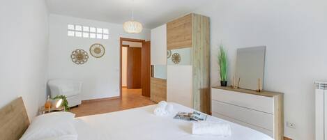A modern hotel room with a large bed, a wooden headboard, a bedside table with a lamp, a mirror, and a dresser.