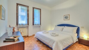 Une chambre à coucher comprenant un lit, une table de chevet, deux fenêtres, un radiateur et un tableau encadré accroché au mur.