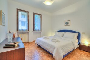 Une chambre à coucher comprenant un lit, une table de chevet, deux fenêtres, un radiateur et un tableau encadré accroché au mur.