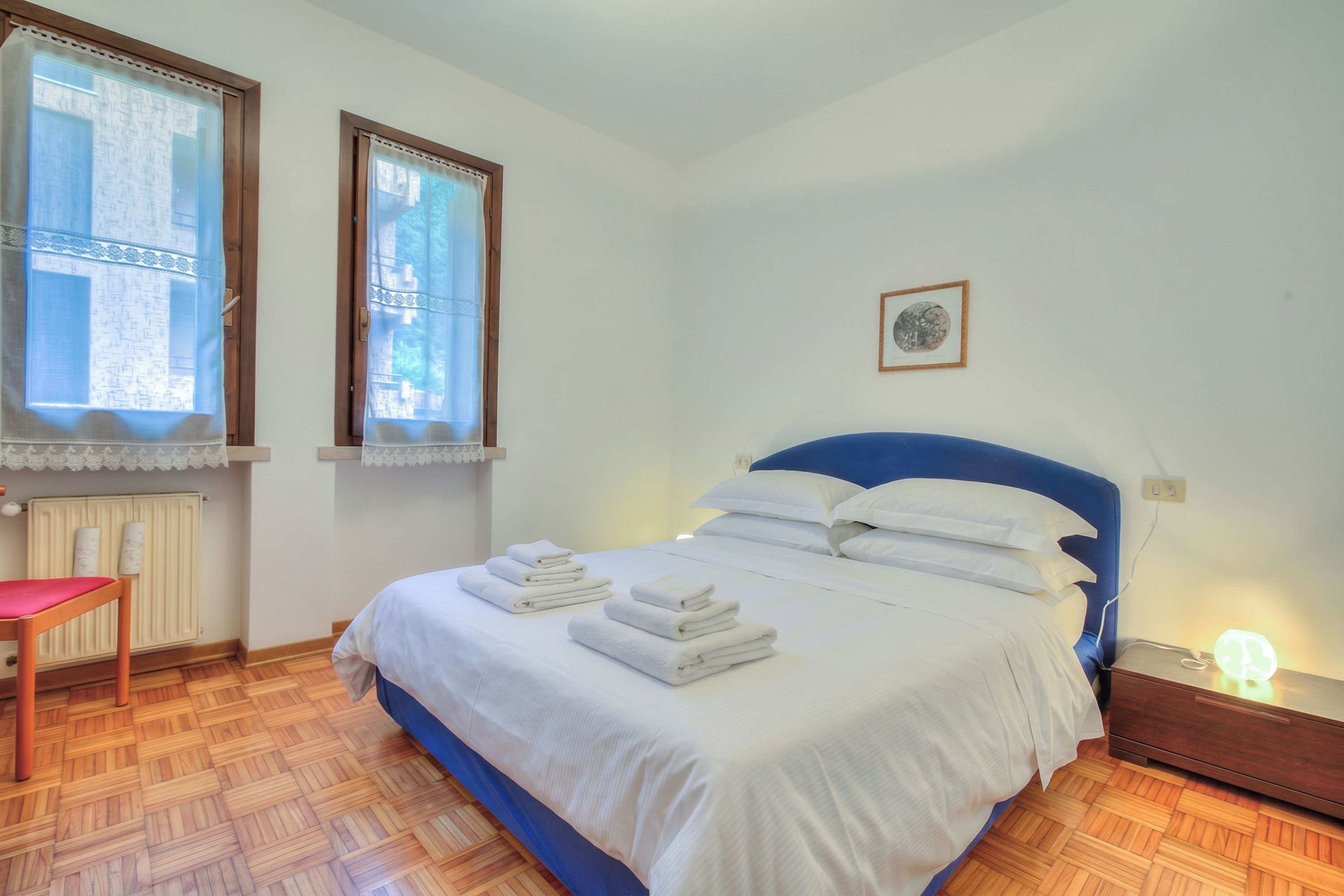 Un dormitori amb un llit, una cadira, una tauleta de nit amb una làmpada i una finestra amb cortines.
