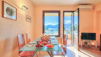 Zona pranzo con tavolo in vetro, sedie, televisione e balcone con vista sulle montagne.