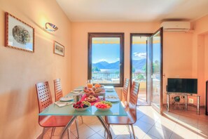 A dining area with a glass table, chairs, a TV, and a balcony with a mountain view. - Appartamento Lake View Laveno 1br, Sleeps 4 (Laveno Mombello)