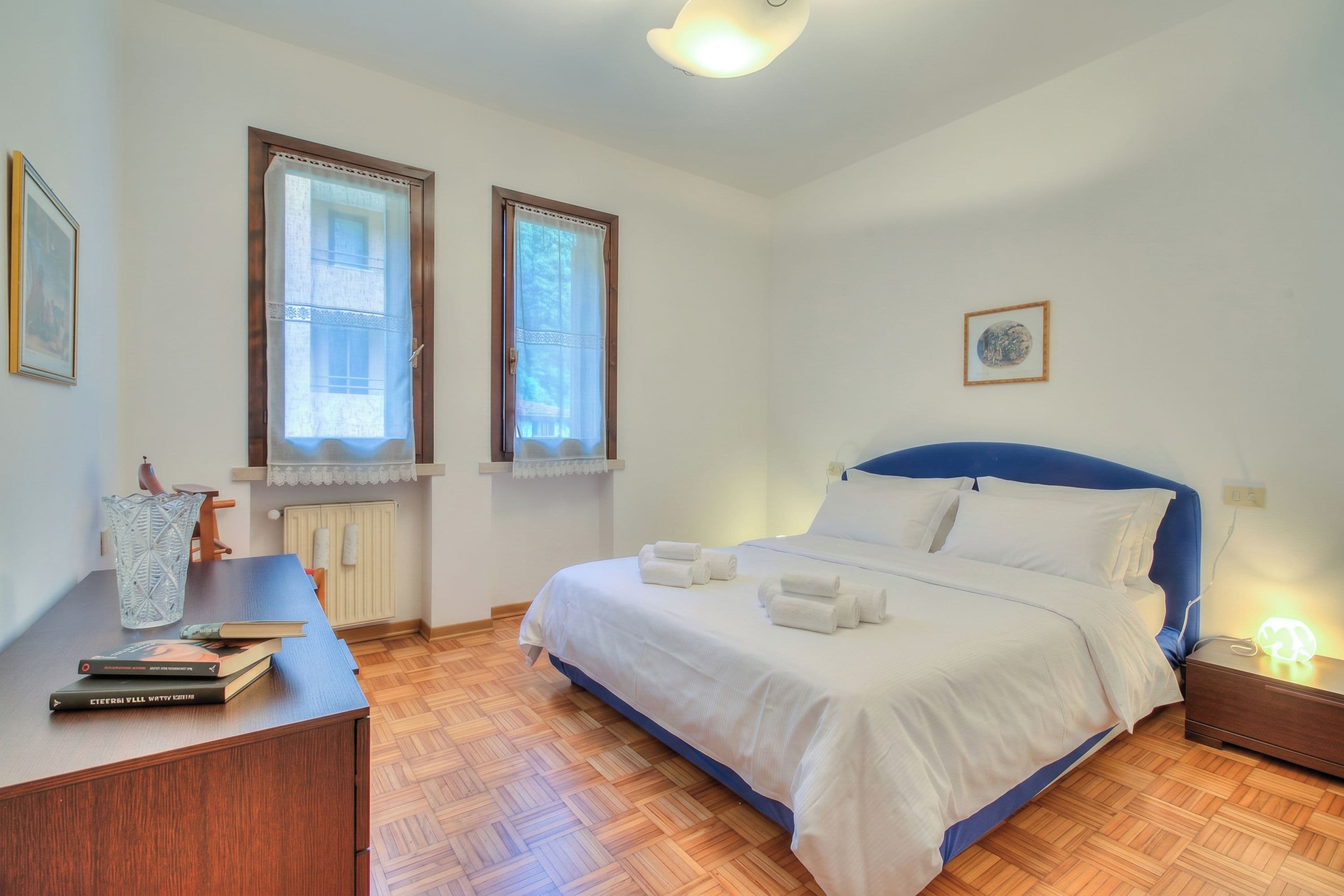 Quarto com cama, mesa de cabeceira, duas janelas, radiador e um quadro na parede.