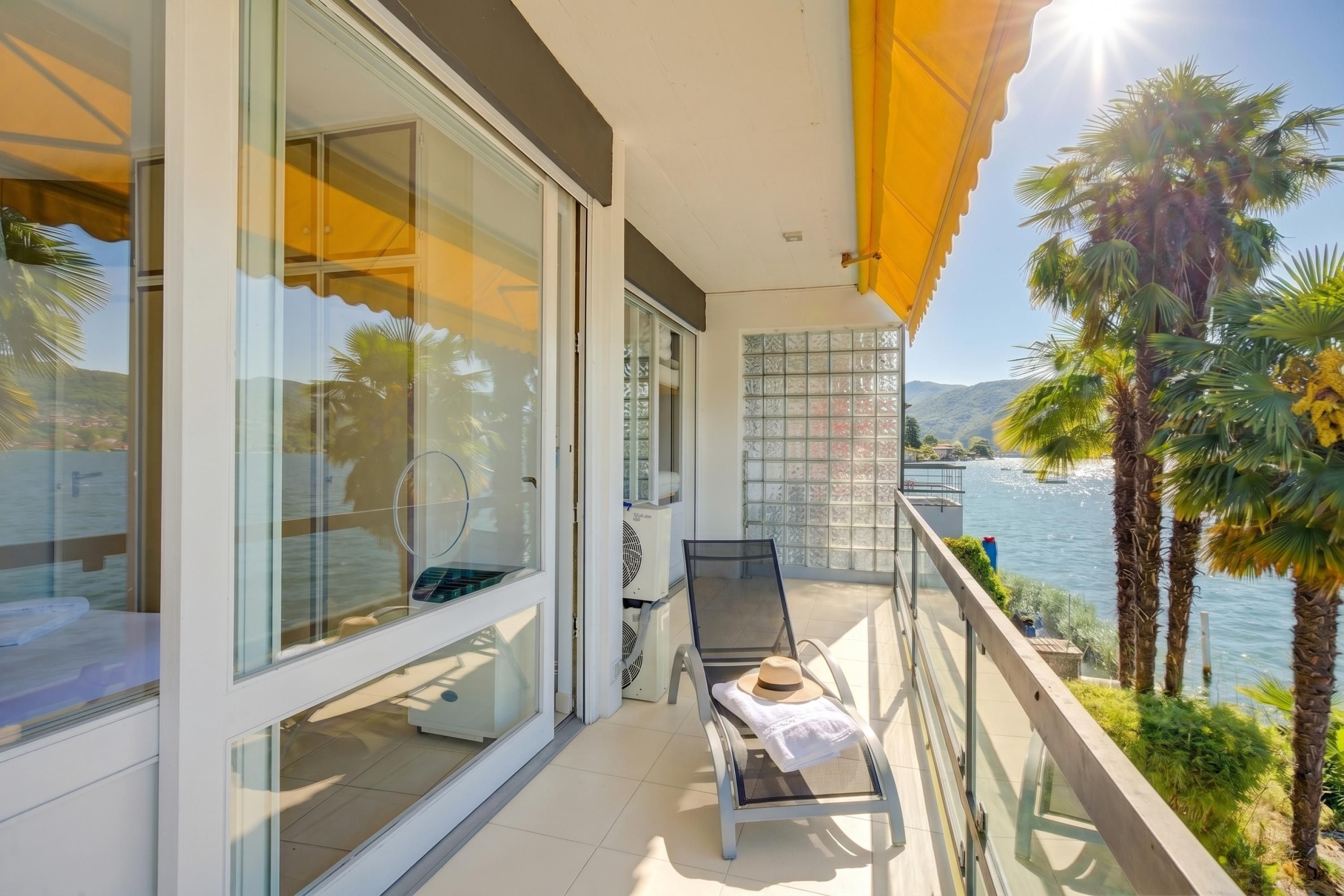 Un balcon avec une porte vitrée, une chaise sur laquelle est posé un chapeau, et une vue sur la mer.