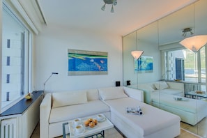 A modern hotel room with a white sofa, a glass coffee table with pastries, a poster of the Australian National Gallery, and a large mirror reflecting the room. - Bellariva On The Lake With View Balcony (Collina d'Oro)