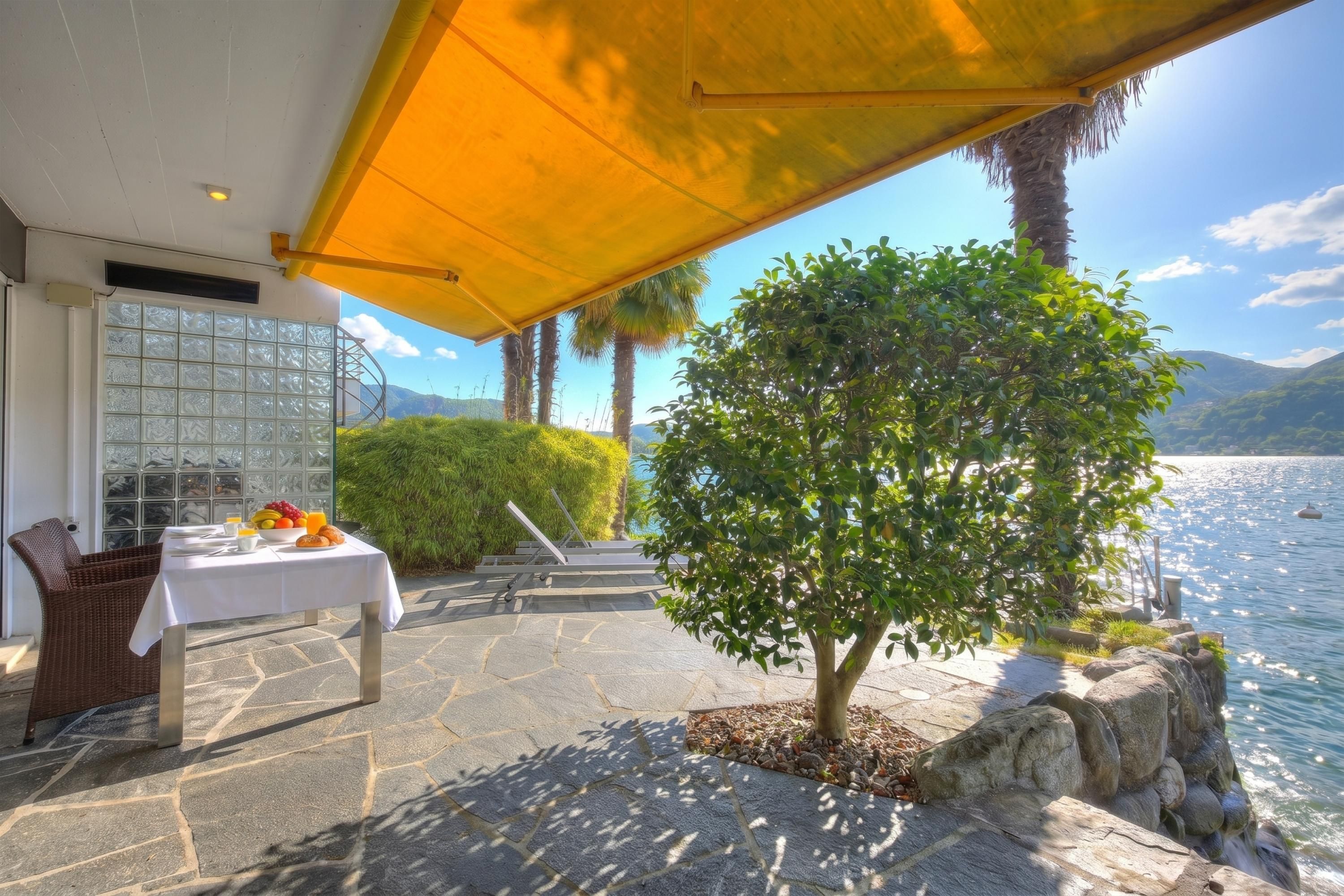 Una veranda con tavolo apparecchiato per mangiare, una sedia in vimini, un albero e vista sull'acqua.