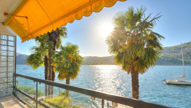 A balcony with a glass railing overlooking a lake, palm trees, and a boat. - Bellariva On The Lake With View Balcony (Collina d'Oro)