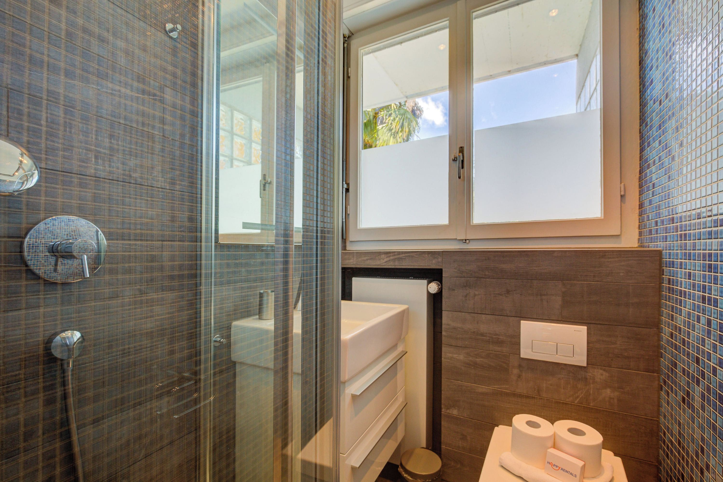 A bathroom with a glass shower enclosure, a toilet with a towel rack, and a window with a view of greenery.