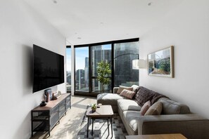 A modern living room with a large flat-screen TV, a sofa, a coffee table, and a view of the city through large windows. - 2BR Skyline Apartment with Pool & Views (Southbank)