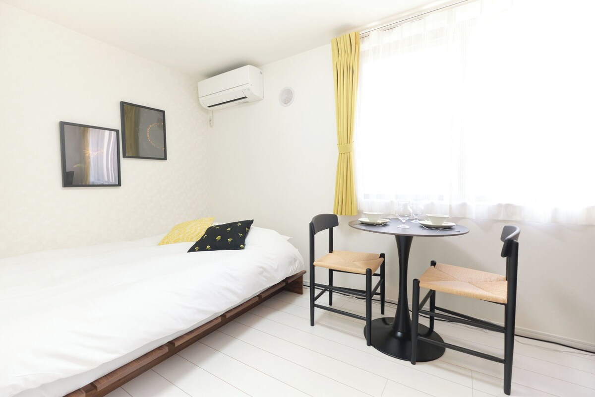 A single bed with a white mattress, a small table with a chair, and an air conditioning unit on the wall.