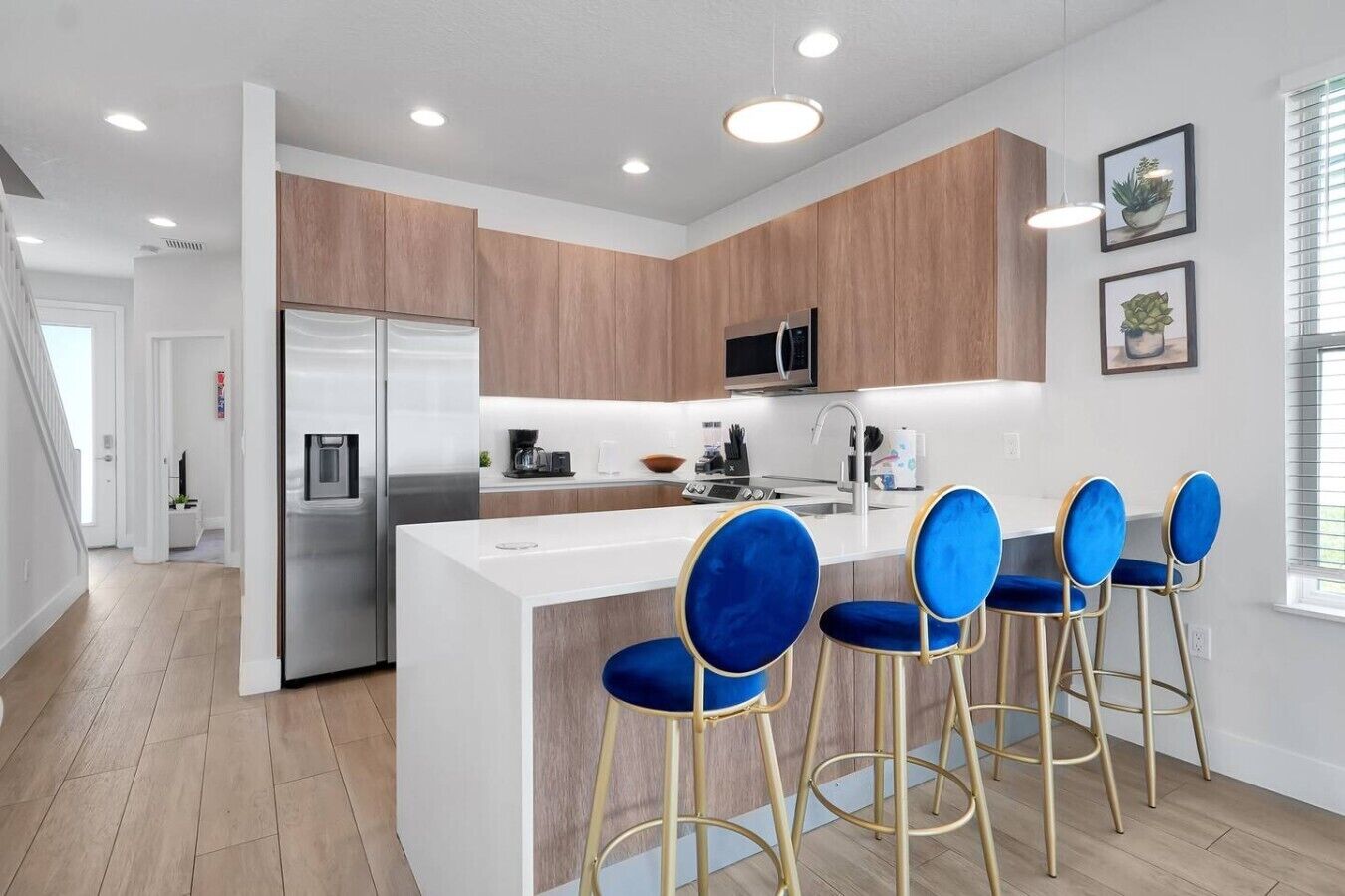 A modern kitchen with wooden cabinets, a stainless steel refrigerator, a microwave, and a bar area with blue bar stools.