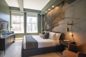 Un dormitorio moderno con una cama grande, un escritorio, una silla y vistas de la ciudad a través de amplios ventanales.