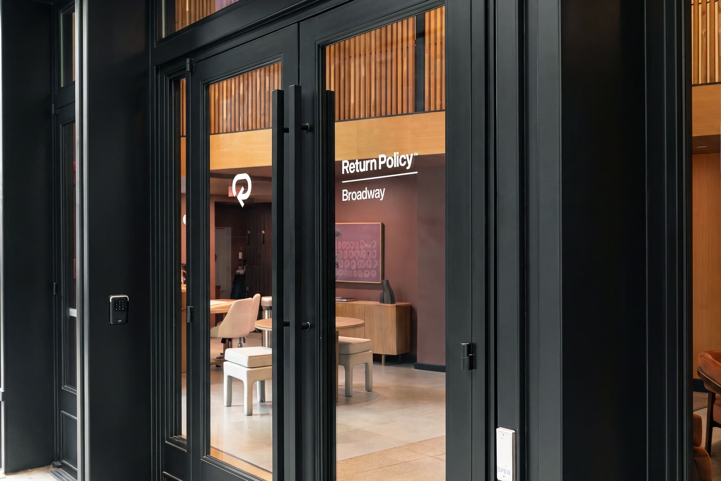 A hotel entrance with a glass door, a sign reading 'Return Policy Broadway,' and an interior view with a reception desk and seating area.