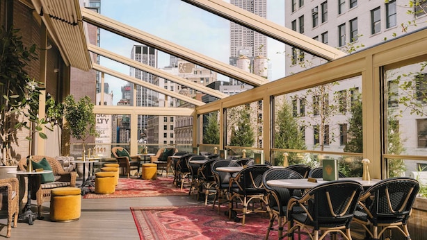 Um restaurante no terraço com mesas ao ar livre, janelas amplas e vista para edifícios da cidade.