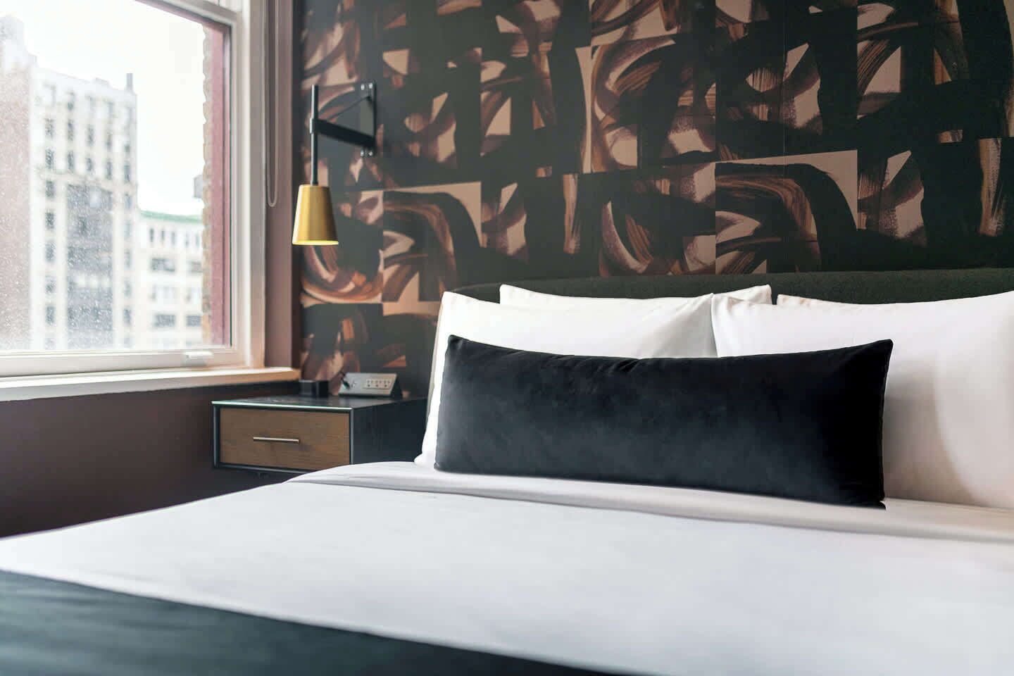 A hotel room with a bed, a nightstand, a lamp, and a window with a view of buildings.