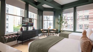 A hotel room with a bed, a desk with a computer, a chair, a small table with a plant, and large windows with a city view. - NoMad Hotel Near Broadway & 5th Ave (New York)