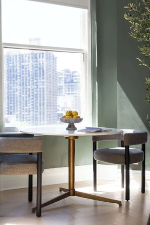 A dining area with a table and chairs, a bowl of lemons, and a view of tall buildings through a window. - NoMad Hotel Near Broadway & 5th Ave (New York)