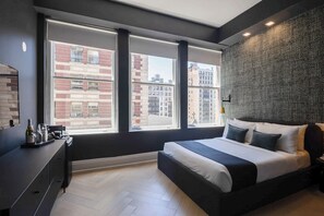 A modern bedroom with a large bed, a dresser, and a view of city buildings through large windows.