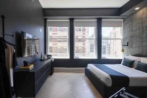 Un dormitorio moderno con cama, cómoda y ventanas amplias con vistas a un paisaje urbano.