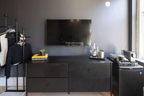A modern room with a flat-screen TV mounted on the wall, a black dresser with drawers, a clothing rack with jackets, and a small potted plant.