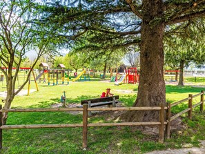 A playground with various play structures, a bench, and a large tree in the foreground. - La Baita dei Nonni by Interhome (Gera Lario)