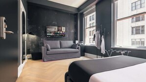 Ein modernes Hotelzimmer mit grauem Sofa, einem Bett, einem Fernseher und Blick auf die Stadt durch große Fenster.