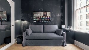 Ein modernes Wohnzimmer mit einem grauen Sofa, einem angebrachten Fernseher, der einen Film zeigt, einer Lampe und Blick auf Gebäude draußen.