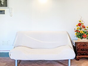 Un canapé blanc dans une pi èce aux murs blancs, une table d’appoint en bois sur laquelle est posé un vase de fleurs, et un sol carrelé.