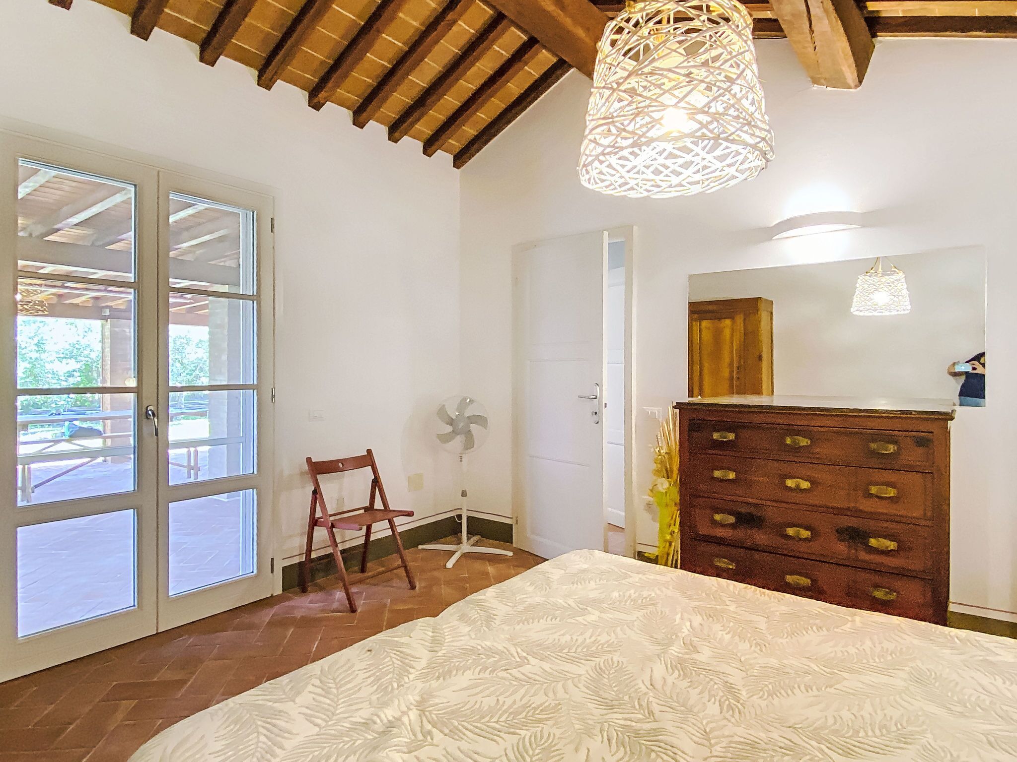 A bedroom with a wooden ceiling, a bed with a patterned bedspread, a dresser with drawers, a chair, and a fan.