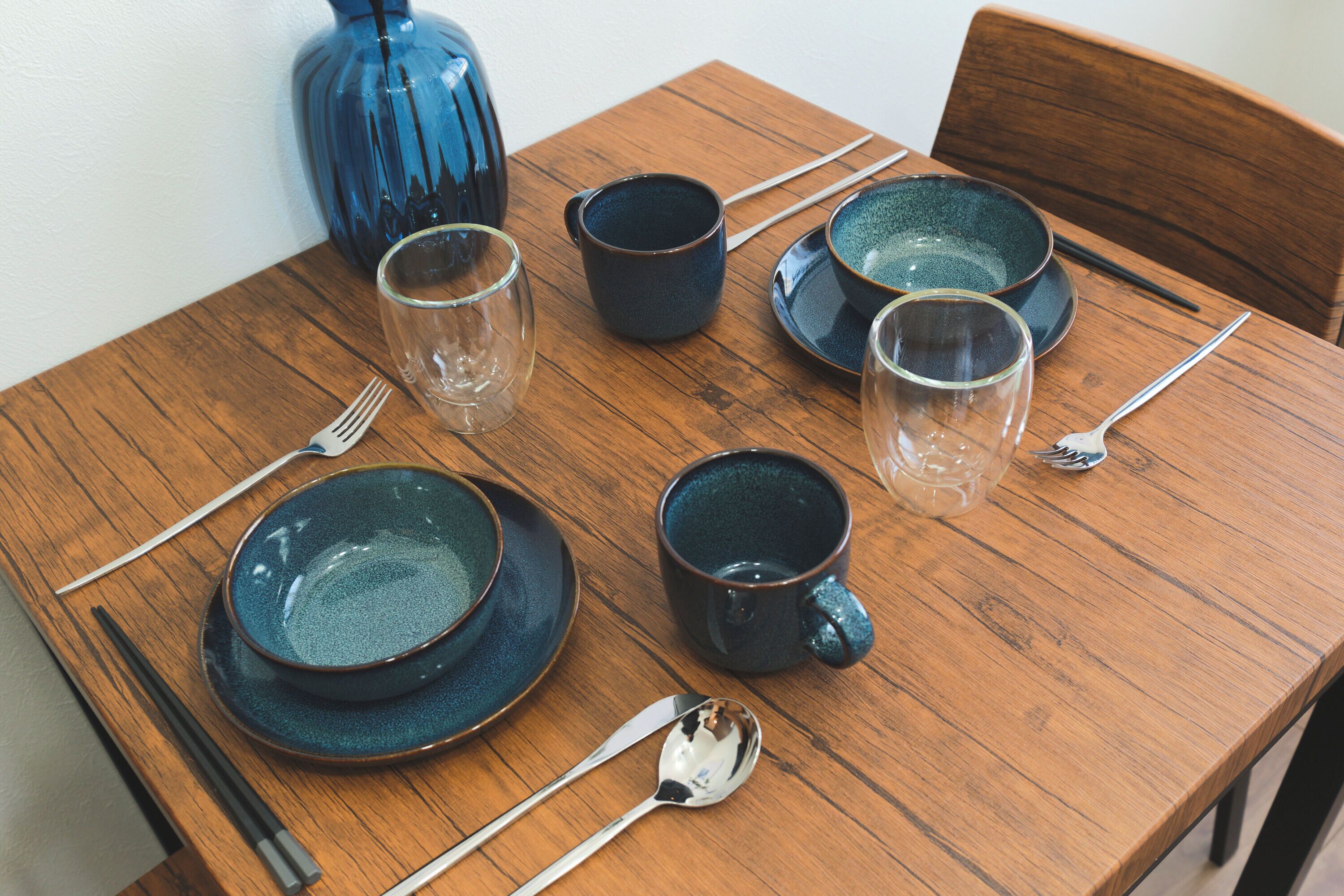 A wooden dining table with ceramic plates, bowls, and cups, accompanied by chopsticks and spoons.