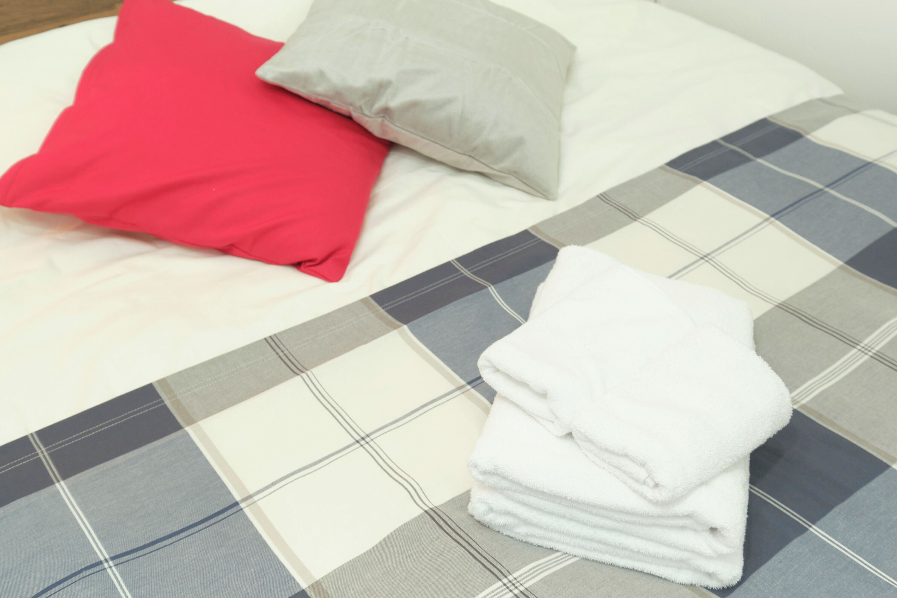 A bed with a plaid bedspread, a red pillow, a gray pillow, and a stack of white towels.