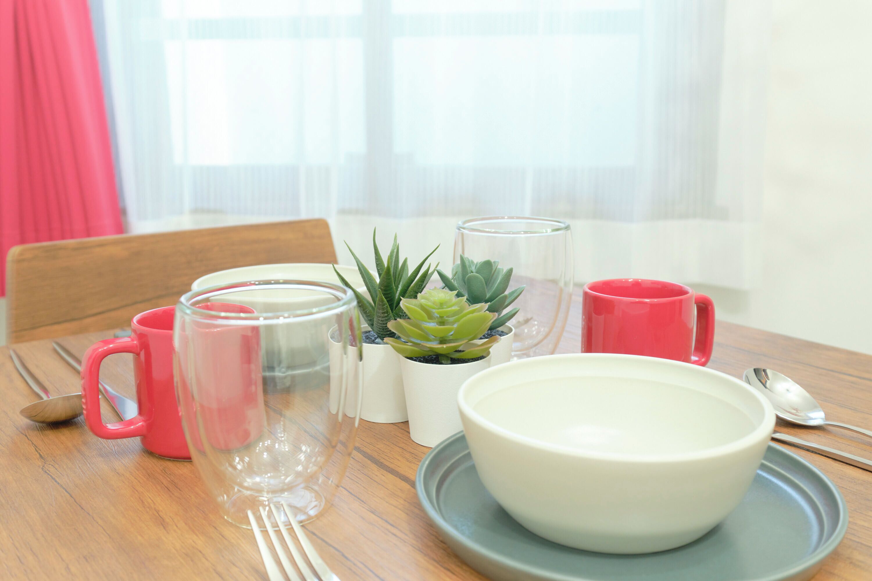 Une table à manger avec un bol blanc, un pichet en verre, une tasse rouge, un verre transparent et une petite plante en pot.