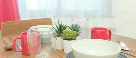 Une table à manger avec un bol blanc, un pichet en verre, une tasse rouge, un verre transparent et une petite plante en pot.