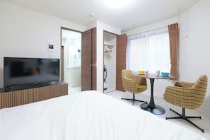 Quarto de hotel com cama, televisão, mesa de jantar com cadeiras e banheiro visível por uma porta aberta.