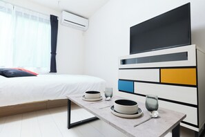 A hotel room with a bed, a table with plates and cups, an air conditioning unit, and a TV on the wall.