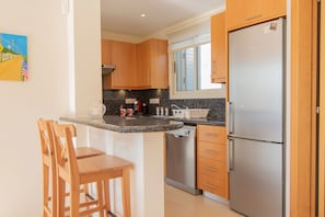 A compact kitchen with wooden cabinets, a stainless steel refrigerator, a dishwasher, and a small bar area with two wooden stools. - Villa Pine View -  a villa that sleeps 6 guests  in 3 bedrooms (Argaka)