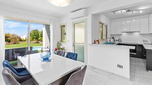Una cocina moderna con armarios blancos, un comedor con mesa y sillas blancas, y vistas a un jardín y piscina al exterior.