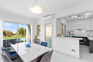 Eine moderne Küche mit weißen Schränken, ein Essbereich mit einem weißen Tisch und Stühlen sowie ein Blick auf Garten und Pool.