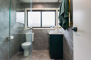 A bathroom with a glass shower enclosure, a toilet, a sink, and a window above the mirror.