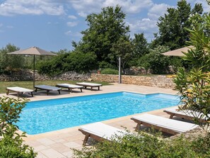 Ein rechteckiger Pool mit Steinrand, umgeben von Liegestühlen und Sonnenschirmen.
