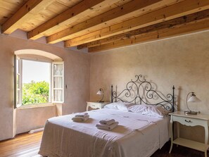 A bedroom with a wooden ceiling, a bed with white linens, two bedside tables with lamps, and a window with a view of greenery. - Vacation home Ita by Interhome (Barbariga)