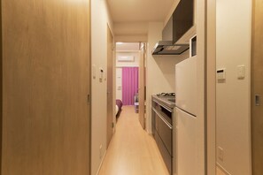 Ein schmales Entree mit einer Holztür links, einer Küchenzeile mit Herd und Backofen rechts und einem Badezimmer mit rosa Duschvorhang im Hintergrund.